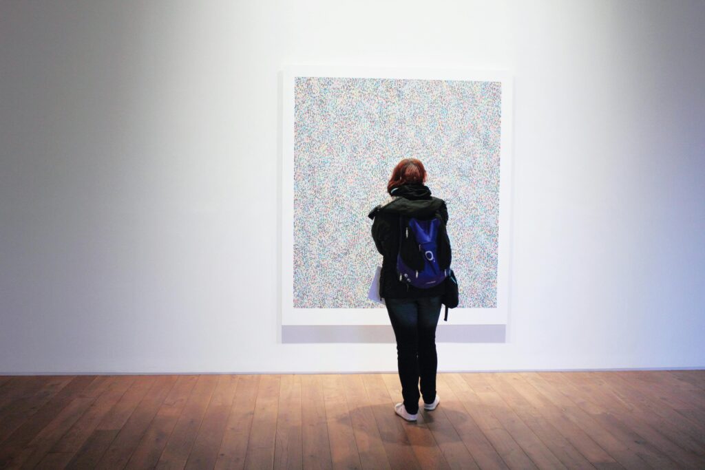 A woman with a backpack observes a colorful painting in an art gallery.